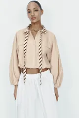 Camisa de popelín color beige con cuello solapa y lazada frontal en contraste con estampado de rayas marrones. Presenta mangas largas abullonadas con puños elásticos, bolsillos con solapa en el pecho y bajo elástico.