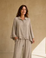 Camisa de lino color beige, de corte oversize y mangas tres cuartos acampanadas con detalles de volados superpuestos en los puños y el ruedo. Presenta cuello camisero y cierre frontal con botones.