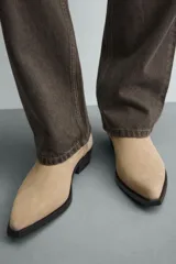 Bota de piel con acabado serraje color beige, con detalle de arnés y hebilla lateral. Presenta horma en punta y suela a contraste con ligero tacón.