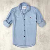 Camisa celeste de manga larga con cuello clásico y cierre frontal con botones negros. Tiene un pequeño bordado de un pájaro rojo en el pecho.