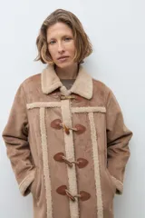 Abrigo de estilo duffle coat en simil gamuza color tostado, con interior y bordes de piel sintética al tono. Presenta cierre frontal mediante alamares de cuero sintético y madera, cuello solapa y mangas largas.