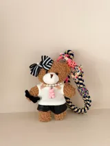 Llavero de peluche con forma de oso, vestido con una remera blanca, shorts negros, un lazo a rayas en la cabeza y un collar de perlas con un dije de osito rosa. Incluye una correa trenzada multicolor con detalles en rosa neón.