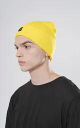Gorro tejido de color amarillo brillante, estilo beanie con borde doblado y parche cuadrado negro con un logo bordado de tijeras cruzadas en blanco.