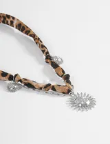 Collar de cinta con estampado animal print, que presenta dijes colgantes en forma de sol en color plateado y un dije central de sol de mayor tamaño. Incluye cierre de mosquetón con cadena de ajuste.