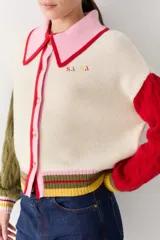 Cardigan de punto con diseño color block, cuerpo en color crema, cuello polo rosado con borde rojo, manga derecha verde oliva y manga izquierda roja. Presenta mangas con tejido de ochos, puños acanalados multicolor y cierre frontal con botones.