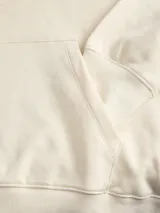 Buzo con capucha y manga larga, de calce holgado, color blanco antiguo (off-white). Presenta un estampado ovalado en la espalda con texto en azul marino que incluye 'Established in Copenhagen 1990', 'Jack&Jones Worldwide', 'Originals Creative Department Daily Basics' y 'From Vision to Reality'.