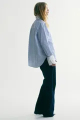 Camisa oversized de popelina de algodón con diseño a rayas verticales en tonos azul y blanco. Presenta cuello clásico y puños anchos en color blanco sólido, cierre frontal con botones, hombros caídos y bajo redondeado con aberturas laterales.