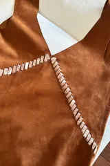 Vestido de gamuza color camel con diseño sin mangas y escote en V. Presenta un detalle decorativo de pasamanería trenzada en el frente que recorre el escote y el centro de la prenda.