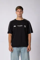 Remera negra de corte regular con cuello redondo y mangas cortas. Presenta detalles de líneas blancas en los hombros y estampado frontal con logo y texto en color blanco.