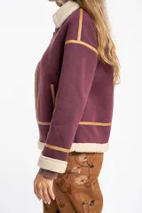 Campera tipo gamulán de color bordó, con interior y cuello de corderito sintético en tono crema. Presenta detalles de ribetes en cuero sintético color suela, cierre frontal metálico y bolsillos laterales.