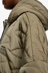 Chaquetón corto de corte línea A, confeccionado en tejido técnico acolchado color beige claro, con costuras en patrón geométrico. Posee cuello subido con capucha integrada, cierre frontal oculto con botones de presión, manga larga con puño elástico y vivos de ribete verde lima al contraste.
