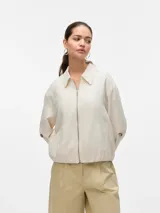 Chaqueta corta color beige, con cuello camisero, cierre frontal de cremallera y bolsillos laterales. Cuenta con mangas largas y detalles de botones en los puños.