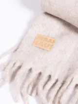 Bufanda de tejido grueso en color gris oscuro, con flecos en los extremos y etiqueta de cuero sintético con el logo de la marca en una de las puntas.