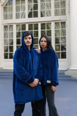 Buzo tipo manta con capucha, confeccionado en tela de peluche suave color azul marino. Presenta un diseño versátil con cordón ajustable en el ruedo para modificar el largo, capucha amplia y un bolsillo frontal de gran tamaño.