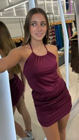 Vestido corto bordó satinado con escote halter fruncido, espalda descubierta y falda ajustada con frunces laterales.