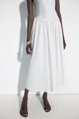 Vestido midi de corte corsetero color blanco, con escote redondo, tirantes finos y detalle de lazo en el delantero. Falda con frunces y cierre en la espalda con cremallera oculta.