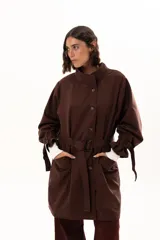 Trench color chocolate con cuello alto, cierre frontal con botones y cinturón ajustable en la cintura. Presenta mangas largas con puños ajustables mediante tiras y hebillas, y bolsillos frontales.