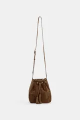 Bolso tipo bandolera de formato mini, confeccionado en gamuza color marrón. Presenta un diseño de cierre fruncido con cordones ajustables y detalle de borlas. Incluye una correa larga ajustable y herrajes metálicos en tono dorado.