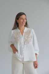 Blusa blanca de corte holgado con cuello mao y cierre frontal de botones. Presenta mangas largas abullonadas con puños elásticos y un delicado bordado en color beige en la parte superior delantera.
