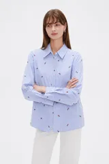Blusa de corte holgado con cuello camisero y cierre frontal de botones. Presenta un diseño de rayas verticales finas en color azul y blanco, decorado con pequeños bordados dispersos.