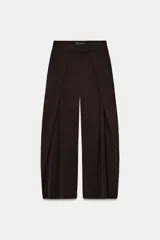 Pantalón de tiro medio con pierna ancha, confeccionado en tela de color marrón oscuro. Presenta pliegues frontales decorativos, bolsillos laterales ocultos en la costura y cierre frontal con cremallera, botón y ganchos metálicos.