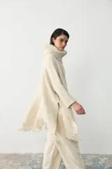 Poncho de tejido artesanal en color crudo, con cuello alto tipo polera, mangas largas y terminaciones con flecos en el ruedo.