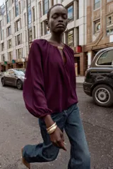 Blusa de tejido semitransparente color violeta, con cuello redondo y detalle de fornitura dorada en el escote. Presenta un diseño de escote en pico, mangas largas abullonadas con puños elásticos y corte holgado.