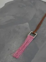 Collar con cordón de cuero tostado y dije geométrico metálico de sección cuadrada con flecos colgantes de hilo rojo.