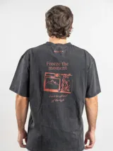 Remera oversize de algodón color negro con efecto lavado. Presenta un estampado gráfico en la espalda con la frase 'Freeze the moment' y un diseño de cámara fotográfica en tonos rojizos.