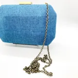 Clutch rígido rectangular forrado en lino azul, con cierre dorado en la parte superior y asa de cadena metálica dorada.