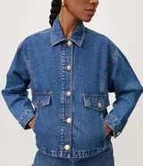 Campera de jean azul de corte recto y holgado, con cuello camisero, cierre frontal de botones metálicos dorados, bolsillos frontales con solapa y mangas largas con puños abotonados.