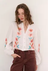 Camisa blanca de manga larga abullonada, con delicados bordados florales en tonos azules y verdes a lo largo de la botonadura frontal y en las mangas. Presenta cuello mao con reborde en contraste y cierre frontal con botones.