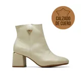 Bota de cuero color beige con taco cuadrado medio y detalle metálico dorado en el talón. Presenta un aplique triangular decorativo con strass en el lateral superior y cierre lateral.