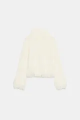 Jersey de punto color blanco crudo, con cuello subido y manga larga. Presenta detalles de flecos en los puños y el dobladillo.