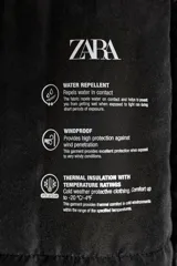 Anorak acolchado negro resistente al agua y al viento, con cuello subido y capucha con pelo sintético en tono tostado a contraste. Cuenta con manga larga, bolsillos delanteros con cierre, cinturón elástico ajustable con hebilla metálica y cierre frontal con cremallera y botones a presión ocultos.
