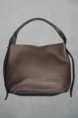 Bolso tipo saco de gran tamaño en cuero marrón oscuro, con textura granulada. Presenta un asa de hombro ancha y detalles metálicos dorados en la unión del asa. Incluye un bolsillo interior extraíble y cierre mediante botón imantado.