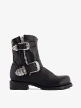 Bota corta de cuero negro, estilo motorista, con caña baja y punta redonda. Presenta cierre lateral de cremallera y tiras ajustables con hebillas metálicas decorativas de estilo western.