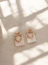 Par de aros colgantes con diseño geométrico compuesto por una pieza superior metálica de forma irregular y una pieza inferior cuadrada de resina con efecto nacarado.