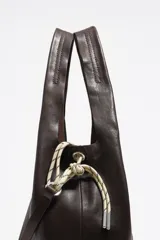 Bolso de mano grande de estructura blanda confeccionado en piel vacuna color marrón oscuro. Presenta asas fijas, cierre de cordón ajustable en los laterales y logo metálico de la marca en la parte frontal.
