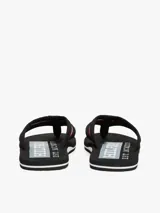 Ojotas negras con suela negra y blanca, y tiras de tejido trenzado con diseño chevron y detalle de cinta distintiva con los colores de la marca.