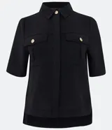 Camisa negra de manga corta con cuello inglés, dos bolsillos delanteros con solapa y botones dorados. Presenta un tejido texturizado y un dobladillo trasero más largo (barra mullet).
