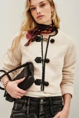 Saco corto de tejido tipo boucle en color crudo, con cierre frontal mediante tres trabas de cuero sintético negro y botones esféricos. Presenta ribetes en contraste negro a lo largo de la abertura frontal y el cuello redondo.