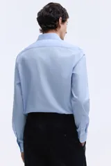 Camisa de tela azul claro con tratamiento de planchado fácil, cuello inglés, tapeta clásica y canesú en la espalda. Mangas largas con tapeta y botón, y cierre ajustable de botón en los puños. Bajo levemente redondeado. Corte ajustado que realza el contorno del cuerpo.