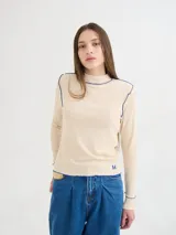 Polera de cuello alto confeccionada en tela rib color beige, con mangas largas y hombreras. Presenta costuras a la vista con pespuntes en hilo azul y un pequeño bordado con la letra M en el ruedo.