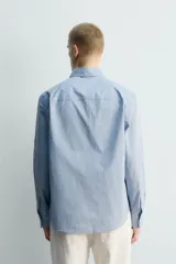 Camisa de hombre de manga larga, corte relajado (relaxed fit), color celeste con patrón de cuadros pequeños en azul y rojo. Presenta cuello abotonado (button-down), bolsillo de parche en el pecho y cierre frontal de botones.