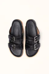 Sandalias de cuero color negro con dos tiras anchas sobre el empeine, cada una con hebillas metálicas ajustables y detalles de tachas plateadas. La suela es de cuero con un borde de color madera.
