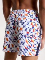 Short de baño blanco con estampado de banderas náuticas en azul, rojo y amarillo. Tiene cintura elástica con cordón ajustable, bolsillos laterales y logo de Tommy Jeans en la pierna.