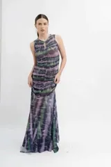 Vestido largo de microtul con estampado tipo batik en tonos azules, grises y blancos. Presenta un diseño sin mangas con cuello redondo y abertura tipo gota en el pecho. El cuerpo está completamente fruncido y forrado, mientras que la falda posee un diseño transparente con terminación en picos que aporta movimiento.