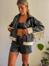 Campera negra de cuero sintético con cierre frontal, detalles en blanco en cuello, puños y ruedo, y flecos blancos en las mangas.