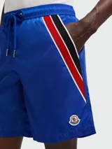 Short de baño azul con cintura elástica y cordón ajustable. Presenta una franja tricolor (rojo, negro y blanco) en el lateral y el logo de la marca en el ruedo.